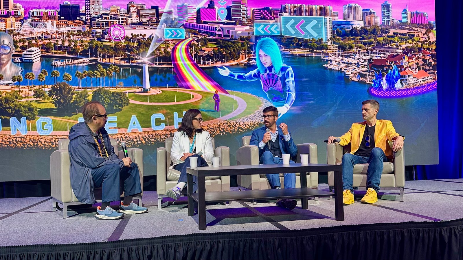 A stage with four XR panelists sitting in armchairs atop a stage at AWE 2025, with Long Beach artwork on the large screen behind them.