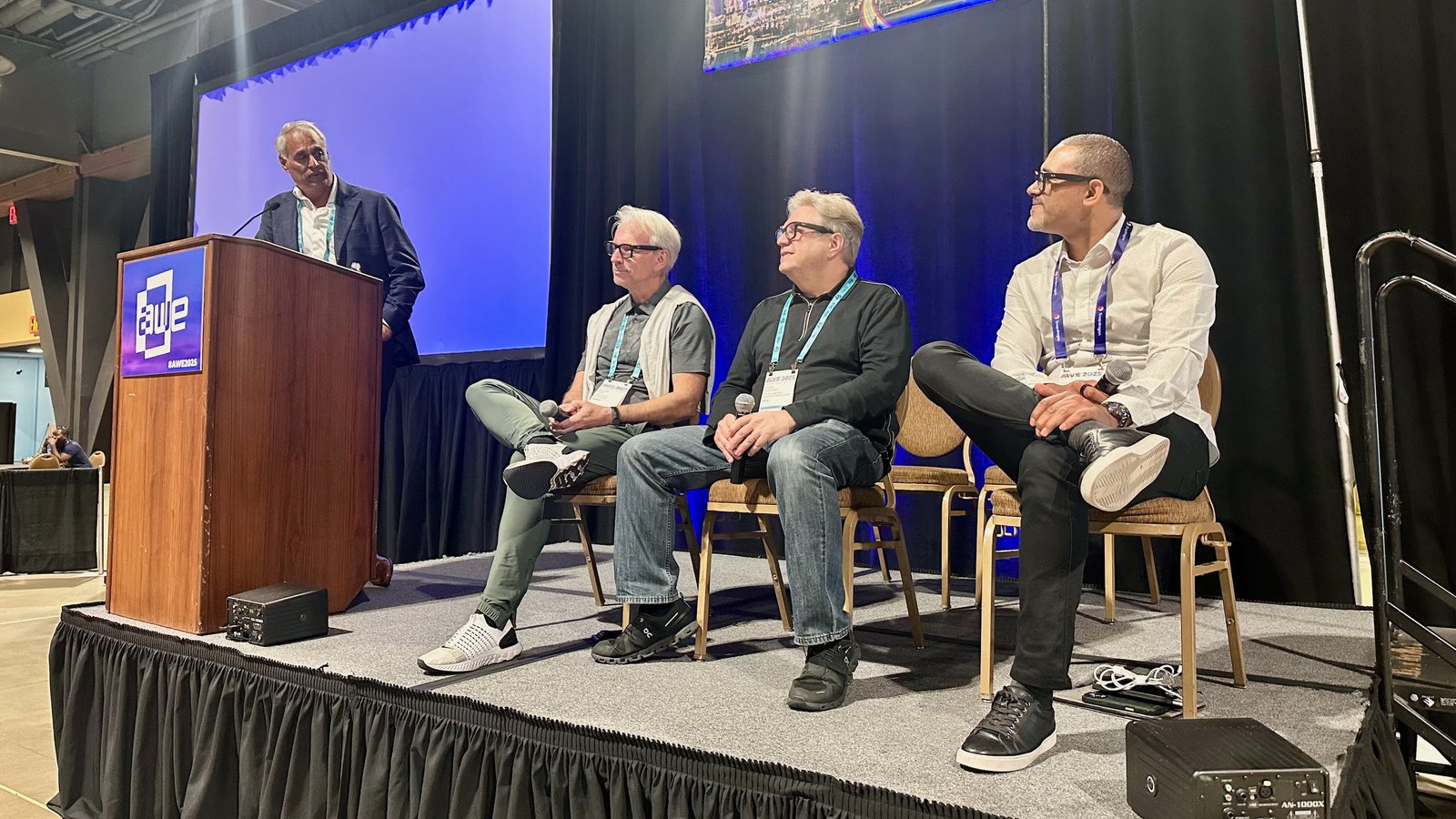 A stage at AWE 2025 with a moderator at the podium and three XR executives from Google, Meta, and Qualcomm seated next to him on stage.