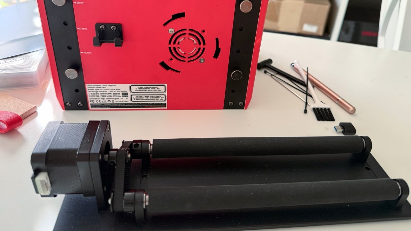 Red laser engraver machine with cylindrical rollers, adjustment knobs, and tools on a table.