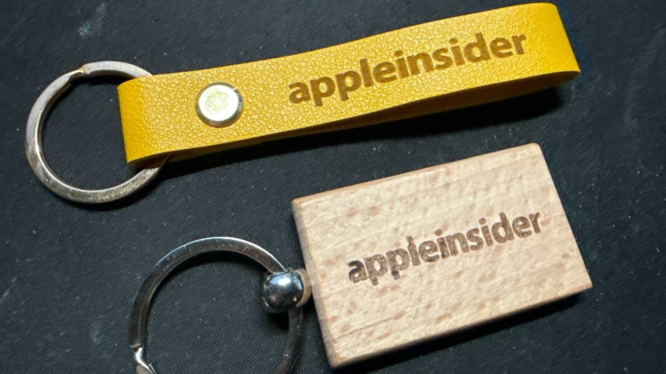 Two keychains on a dark surface, one yellow and leather, the other wooden, both engraved with 'appleinsider'.