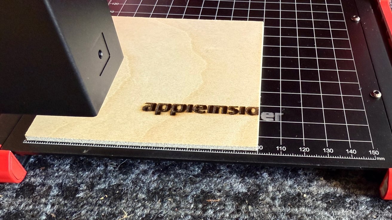 Laser engraver etching the word 'appleinsider' on a wooden surface placed on a black grid mat with red clamps.