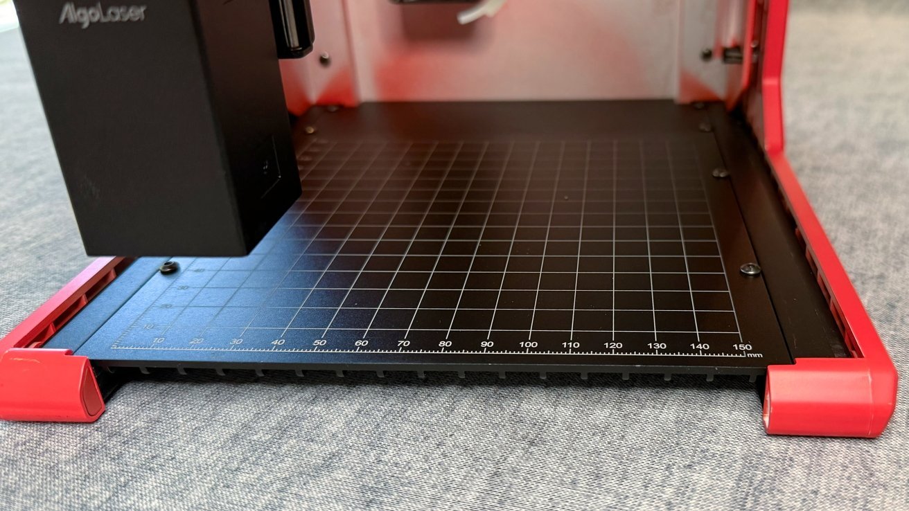 Laser engraver with a black grid base on a fabric surface, featuring a red frame and detailed measurement markings in millimeters along the edge.